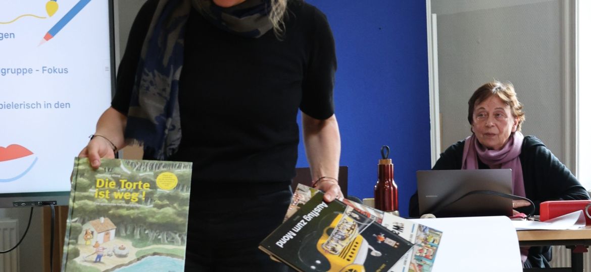 Eine Person zeigt Bilderbücher, eine andere Person sitzt am Schreibtisch im Hintergrund