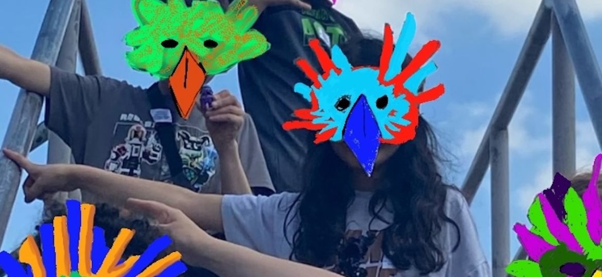 Im Hintergund sieht man den Hafen. Vorne stehen 5 Kinder. Sie tragen bunte Vogelmasken.