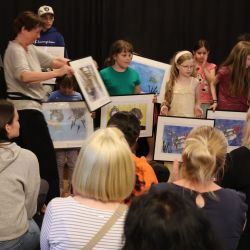 Eine Frau überreicht einem Kind ein Bild. Viel Kinder stehen auf der Bühne.