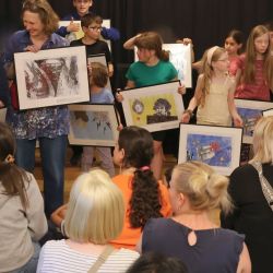 Viele Kinder und eine Frau stehen auf der Bühne und halten Bilder.